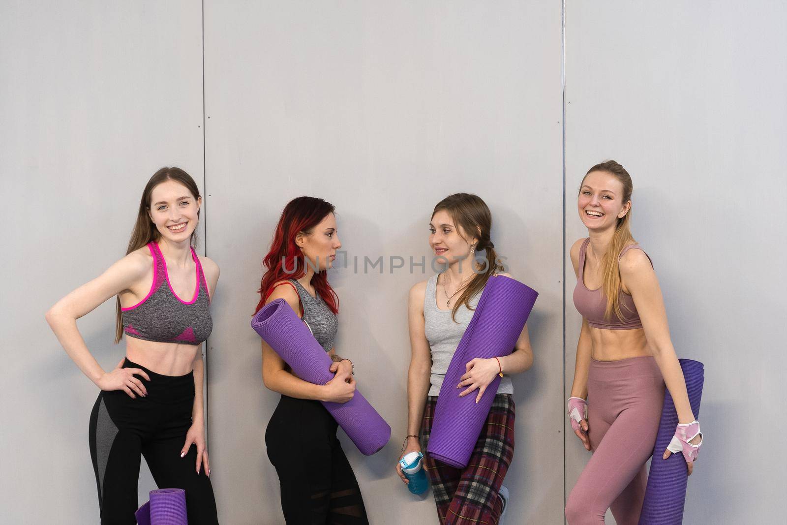 Athletic girls in sports out fits standing next to the wall holding a yoga mat, smiling on camera. Practicing in fitness or yoga studio, working out indoor. Isolated on grey background by LipikStockMedia