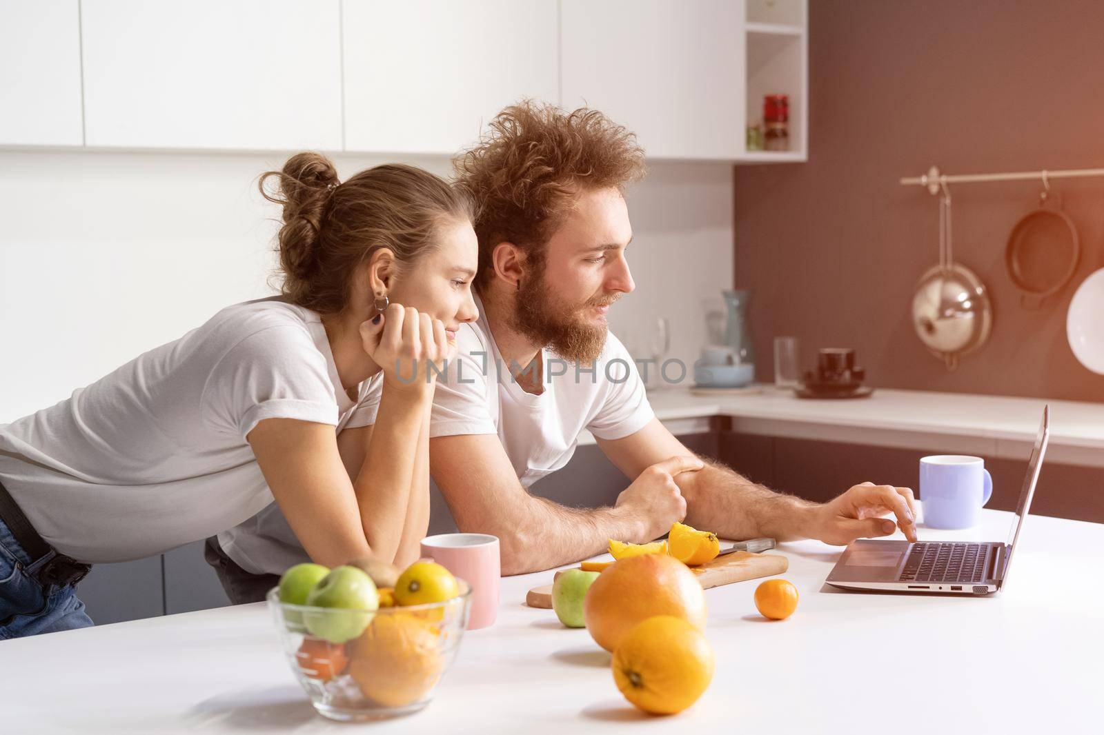 Leaned on kitchen table couple watching romantic movie or streaming social media video. Beautiful young couple talking on video call using laptop. Young couple in kitchen at new home by LipikStockMedia