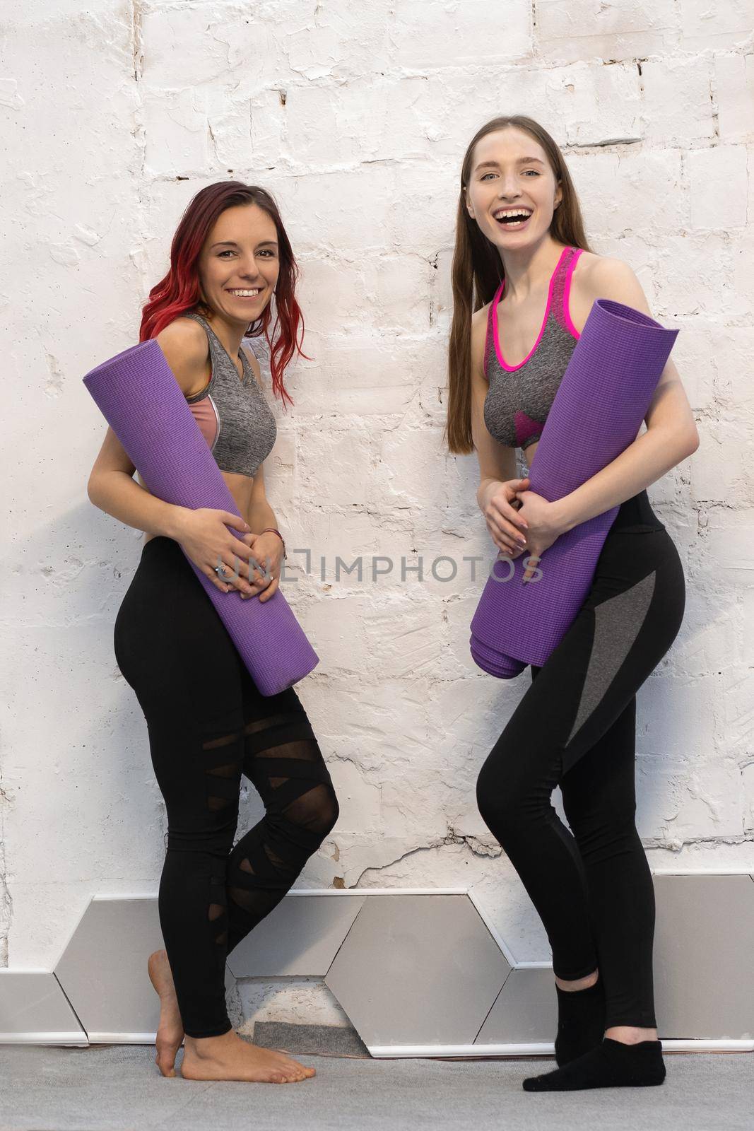 Two pretty fitness girls in sports out fits holding a yoga mat standing next to the white wall, smiling on camera isolated on wall background. Practicing in studio, working out indoor. Loft interior.