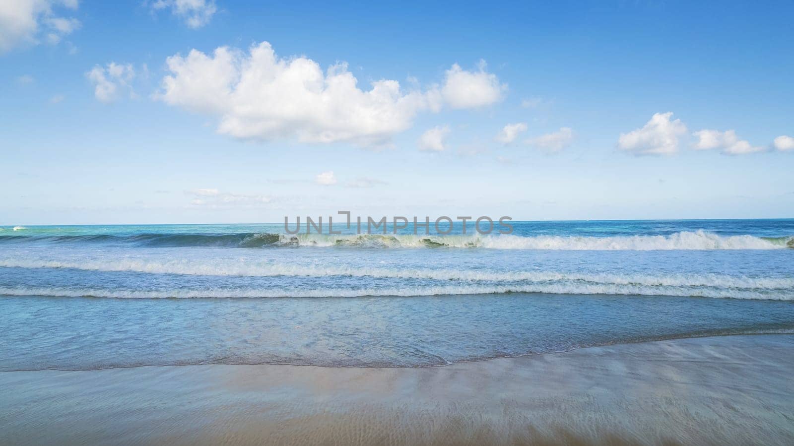 Golden sand and blue ocean water with white clouds. Karon Beach. In places, high waves and splashes. Blue sky. The sun's rays are reflected on the sand. White foam from the waves. Thailand