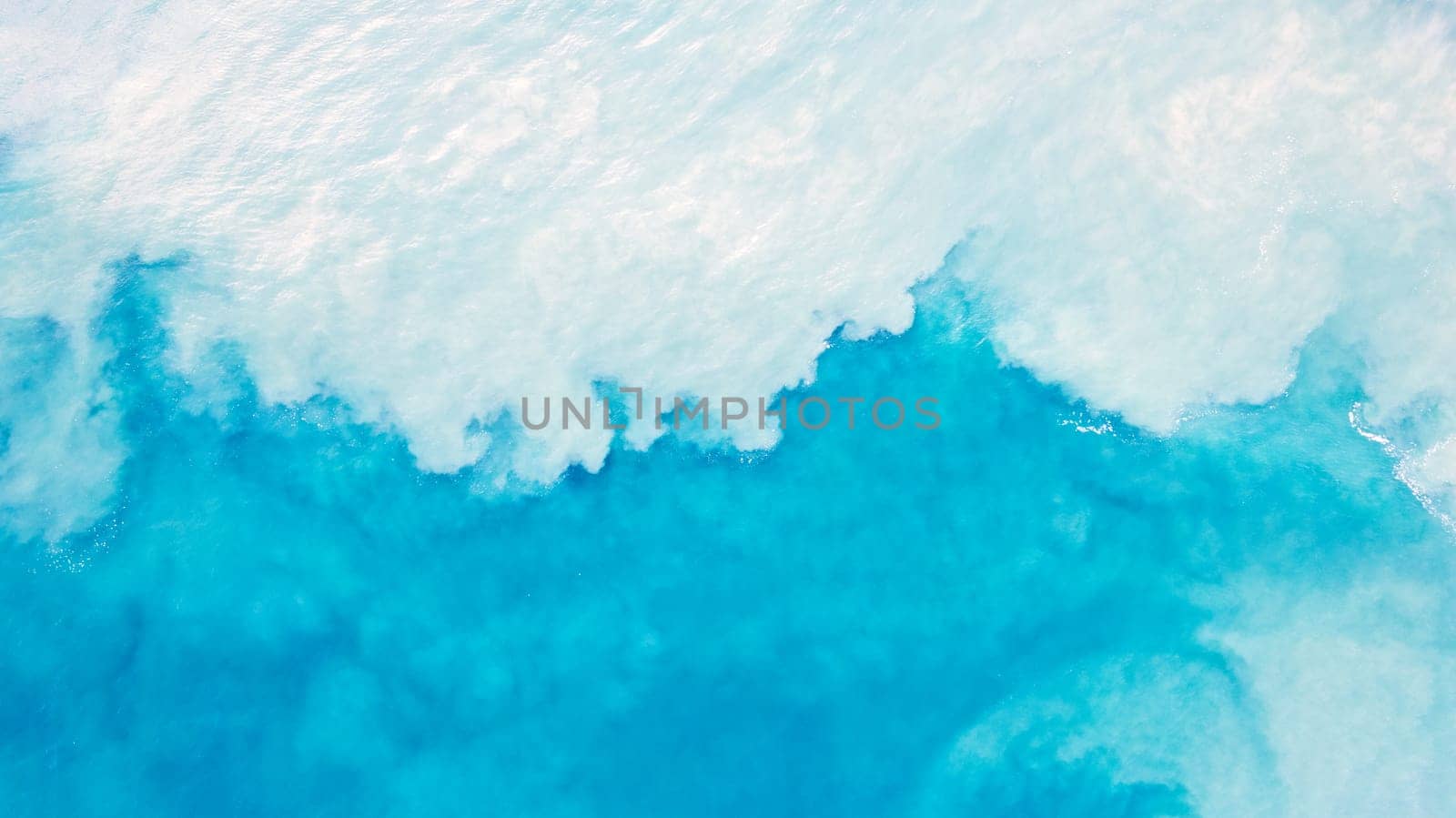 Top view of light blue water with white sand. Waves raise sand and a mix with turquoise water is obtained. Shadows from clouds on a clear ocean. Clear sea. Gradient of blue. The background photo