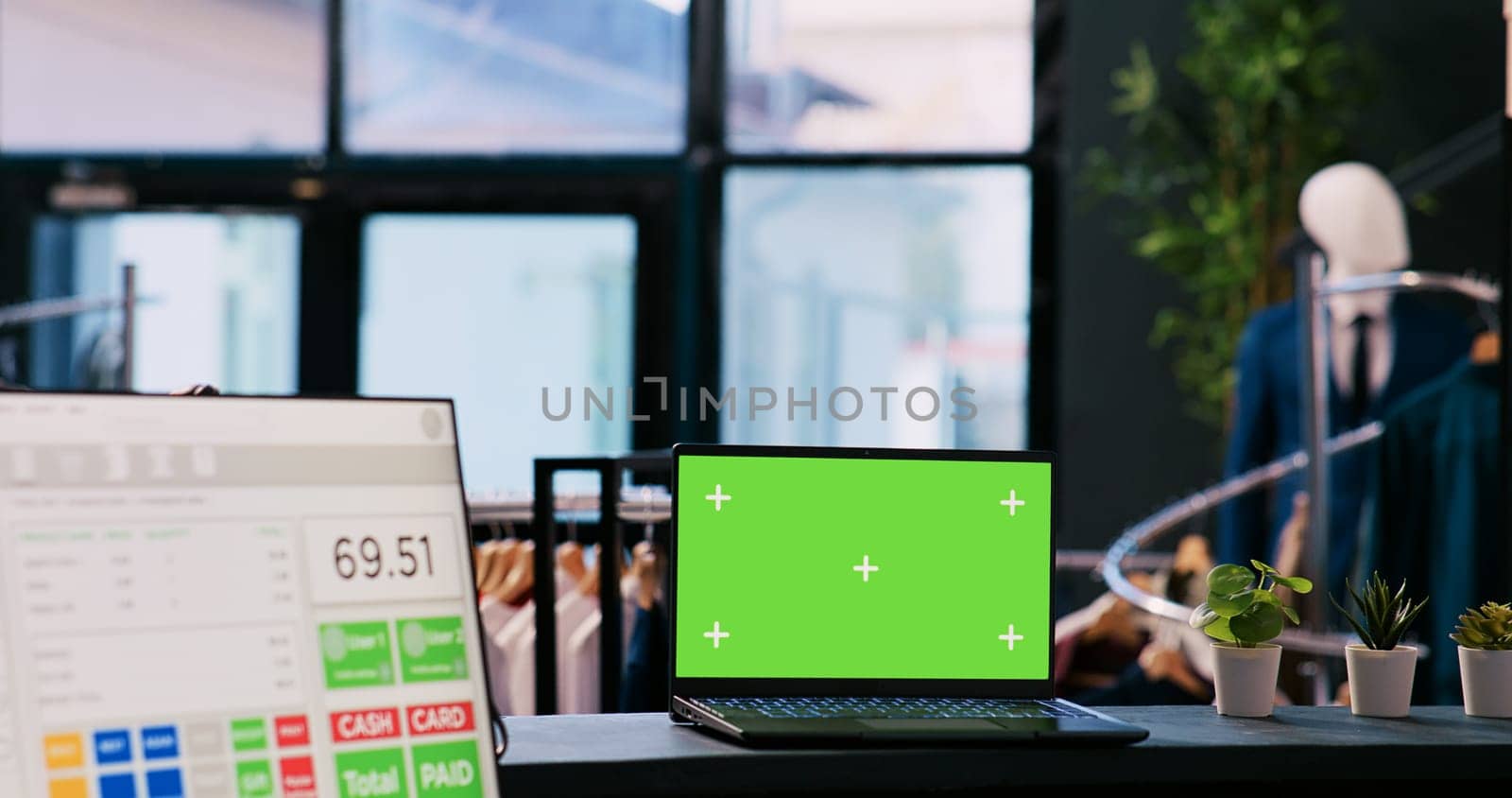 Laptop computer with chroma key green screen mock up display standing on counter desk in modern boutique. In background diverse customers shopping for stylish merchandise in shopping centre