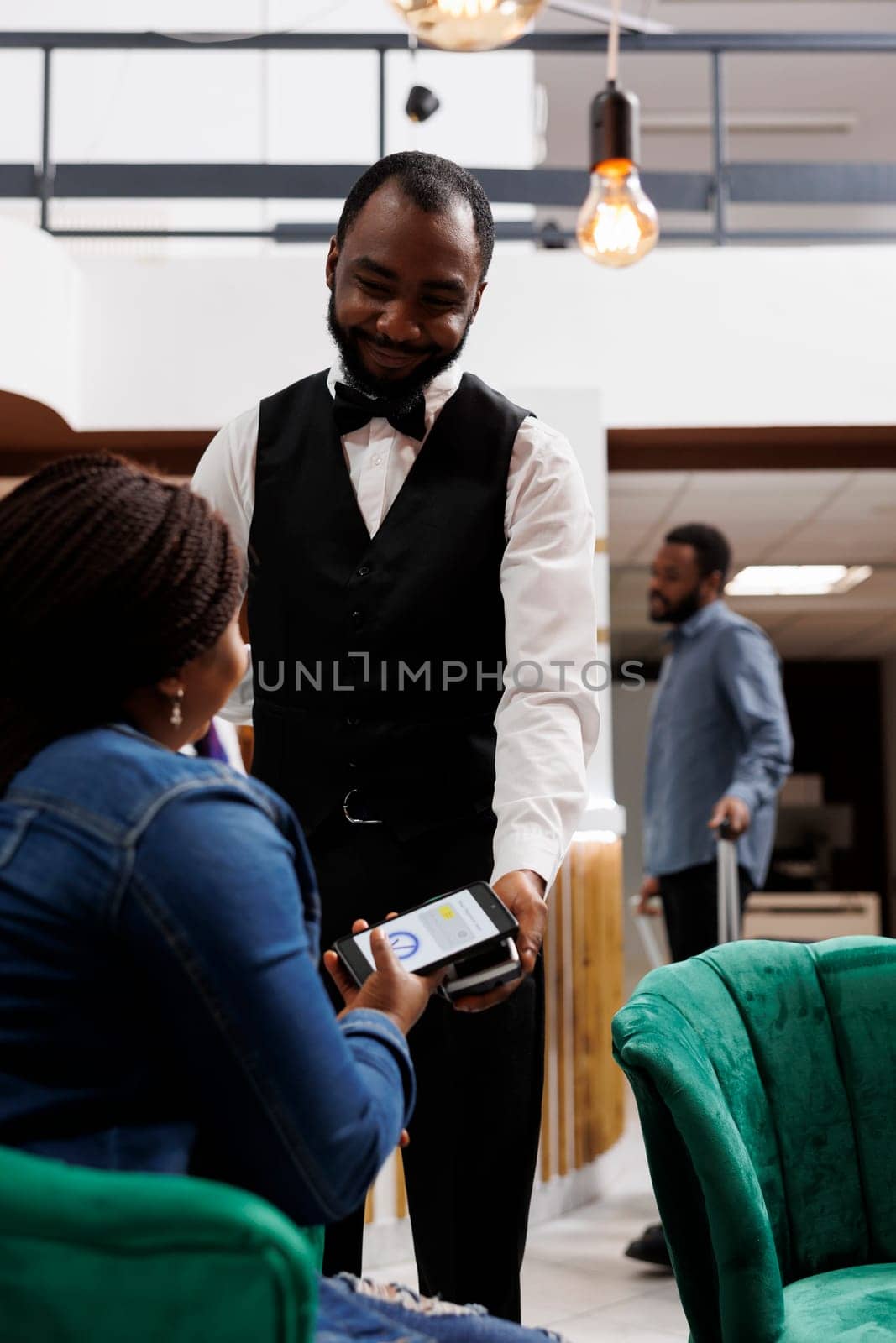Smiling African American man waiter giving pos machine to female hotel guest sitting in lobby. Tourist holding smartphone paying for order while waiting for check-in procedure at lounge area