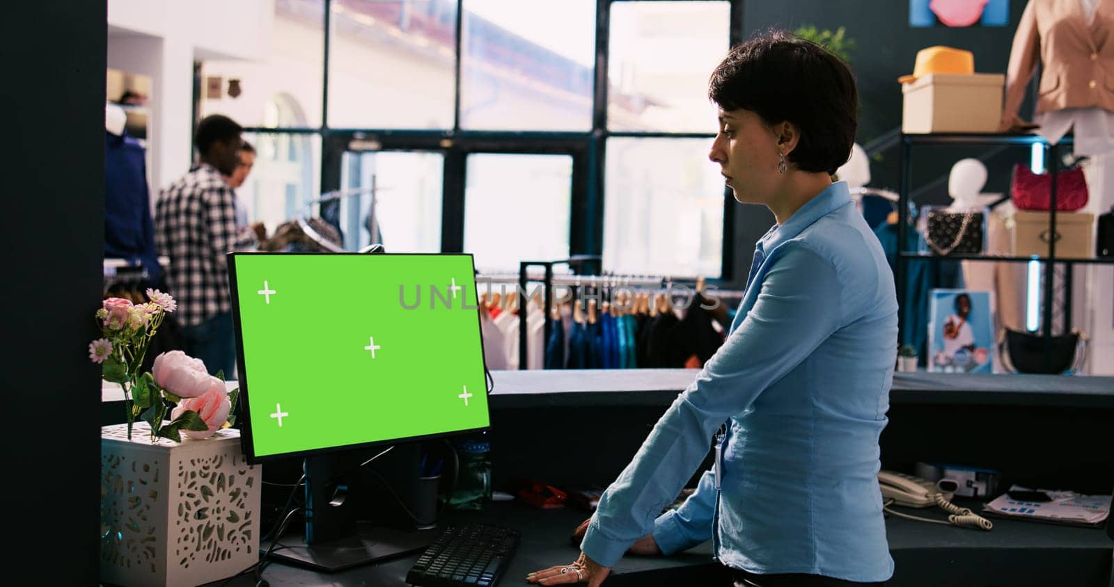 Manager looking at computer with chroma key green screen mock up display, standing at counter desk in modern boutique. Employee waiting for customers in clothing store. Fashion concept