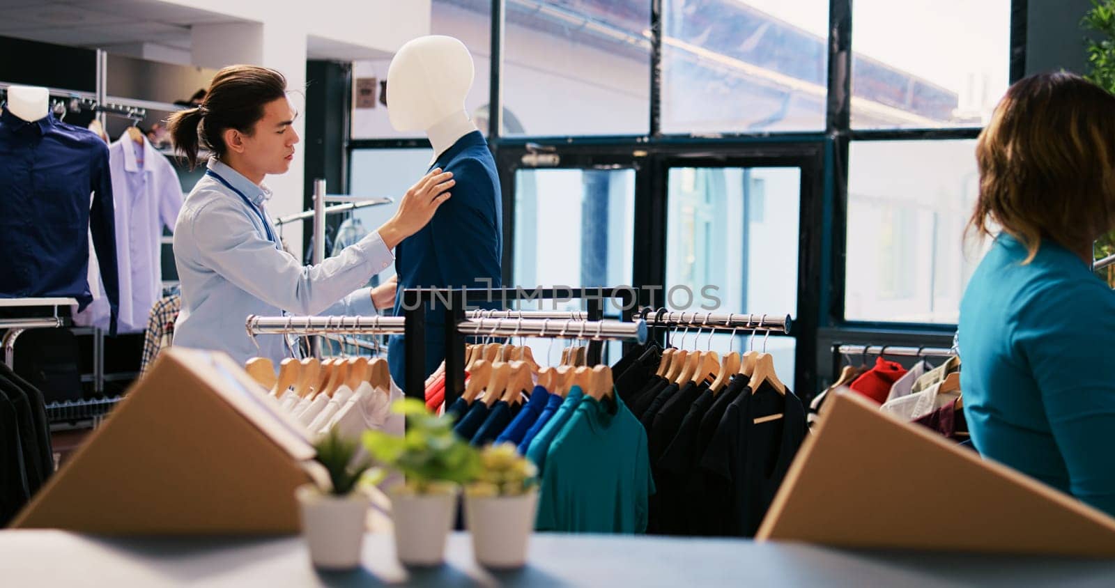 Asian manager closing blue suit buttons, arranging fashionable clothes and stylish accessories in modern boutique. Cheerful employee working at clothing store visual. Fashion concept