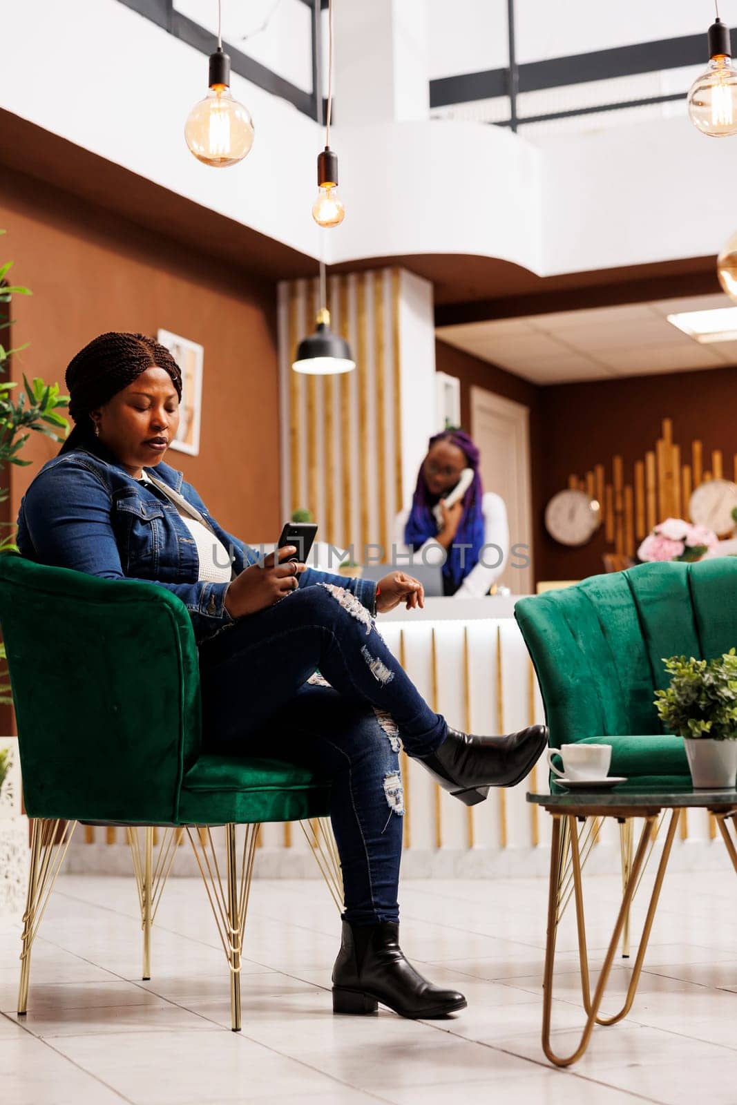 African American woman tourist sitting at hotel lobby using smartphone, connecting wifi, female traveler holding phone browsing internet while waiting for check-in. Mobile technology in hospitality