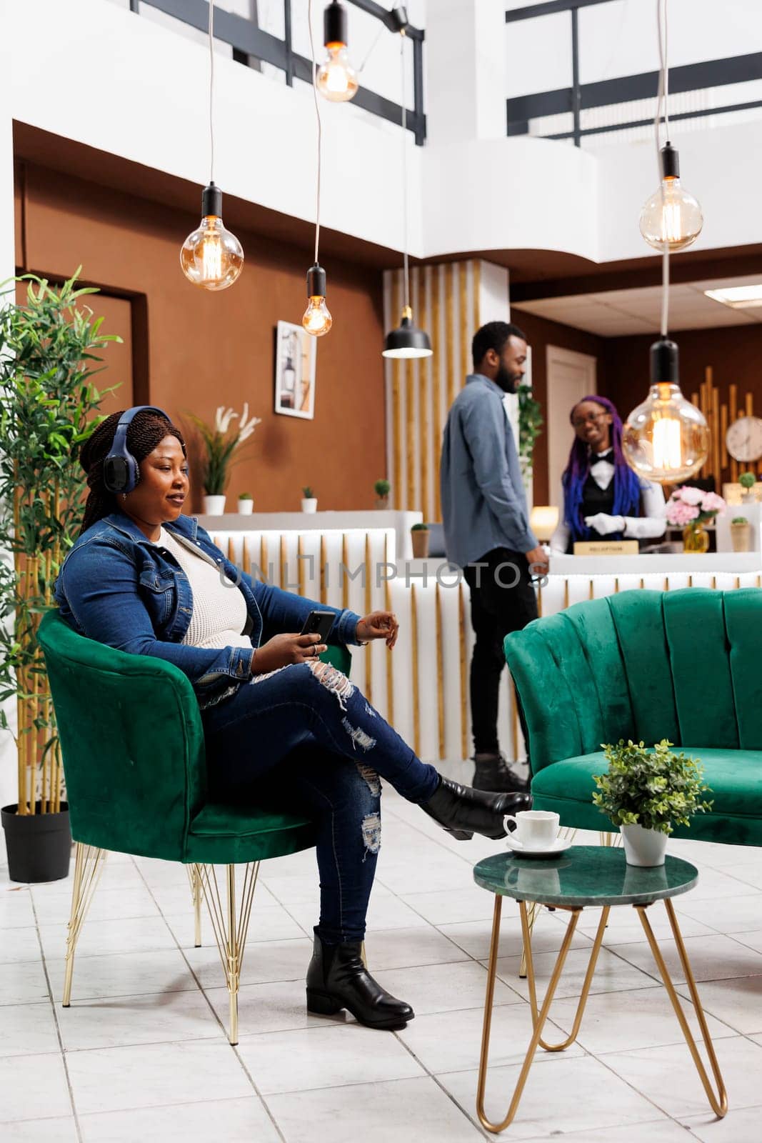 Relaxed African American woman hotel guest sitting in lobby listening music, use wireless headphones and smartphone. Female tourist resting at reception area, waiting for check-in