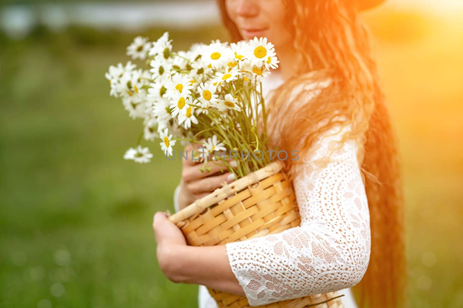 A middle-aged woman in a white dress and brown hat holds a lA middle-aged woman in a white dress and brown hat holds a basket in her hands with a large bouquet of daisies.arge bouquet of daisies in her hands. Wildflowers for congratulations.