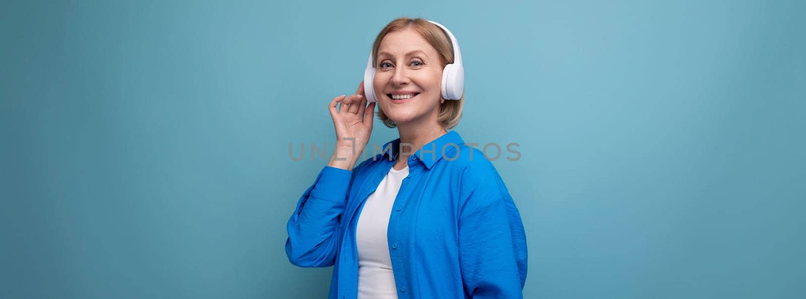 middle age business concept. mature woman listening to audiobook with headphones on blue background copy space by TRMK