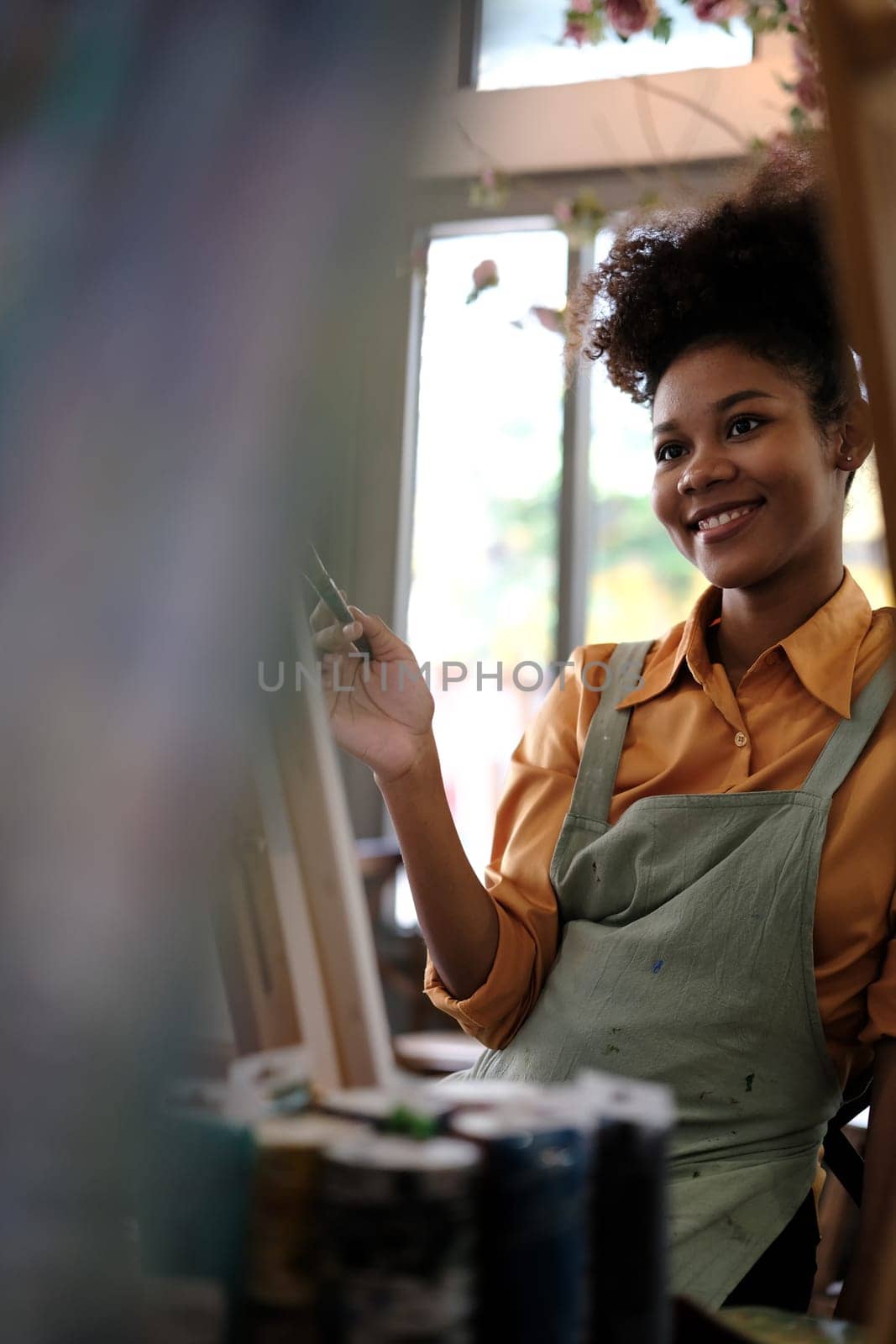 Delighted young woman with Afro haired wearing apron painting picture with brush on canvas in art studio by prathanchorruangsak