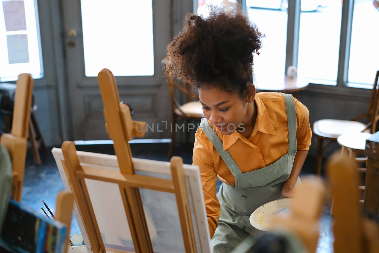 African American female artist wearing apron painting picture with brush on easel in art studio.