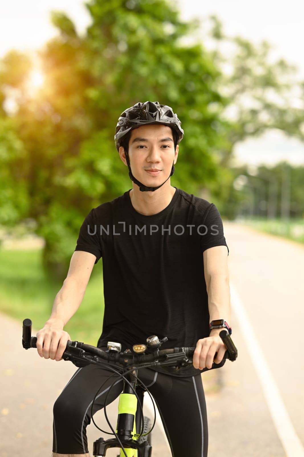 Smiling young asian man dressed in casual clothes and helmet riding a bicycle along the bike path in a city park.