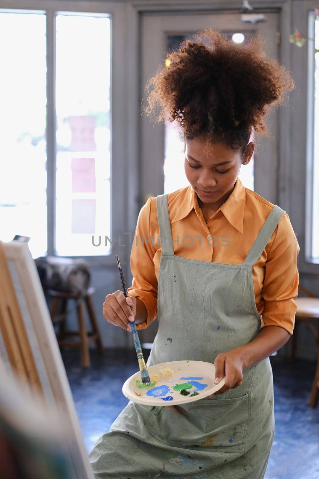 Happy young woman with Afro haired wearing apron painting picture with brush on canvas in art studio.