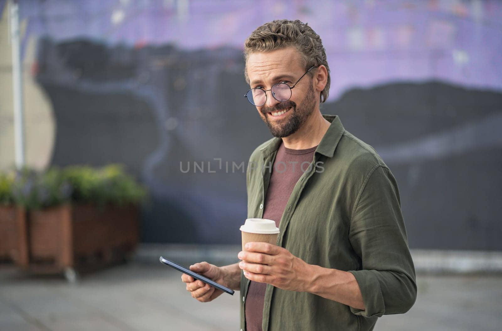 Sarcastic man typing a message on his phone while enjoying a cup of coffee outdoors. With a hint of irony and sarcasm in his expression, he combines technology and leisure in a playful manner. by LipikStockMedia
