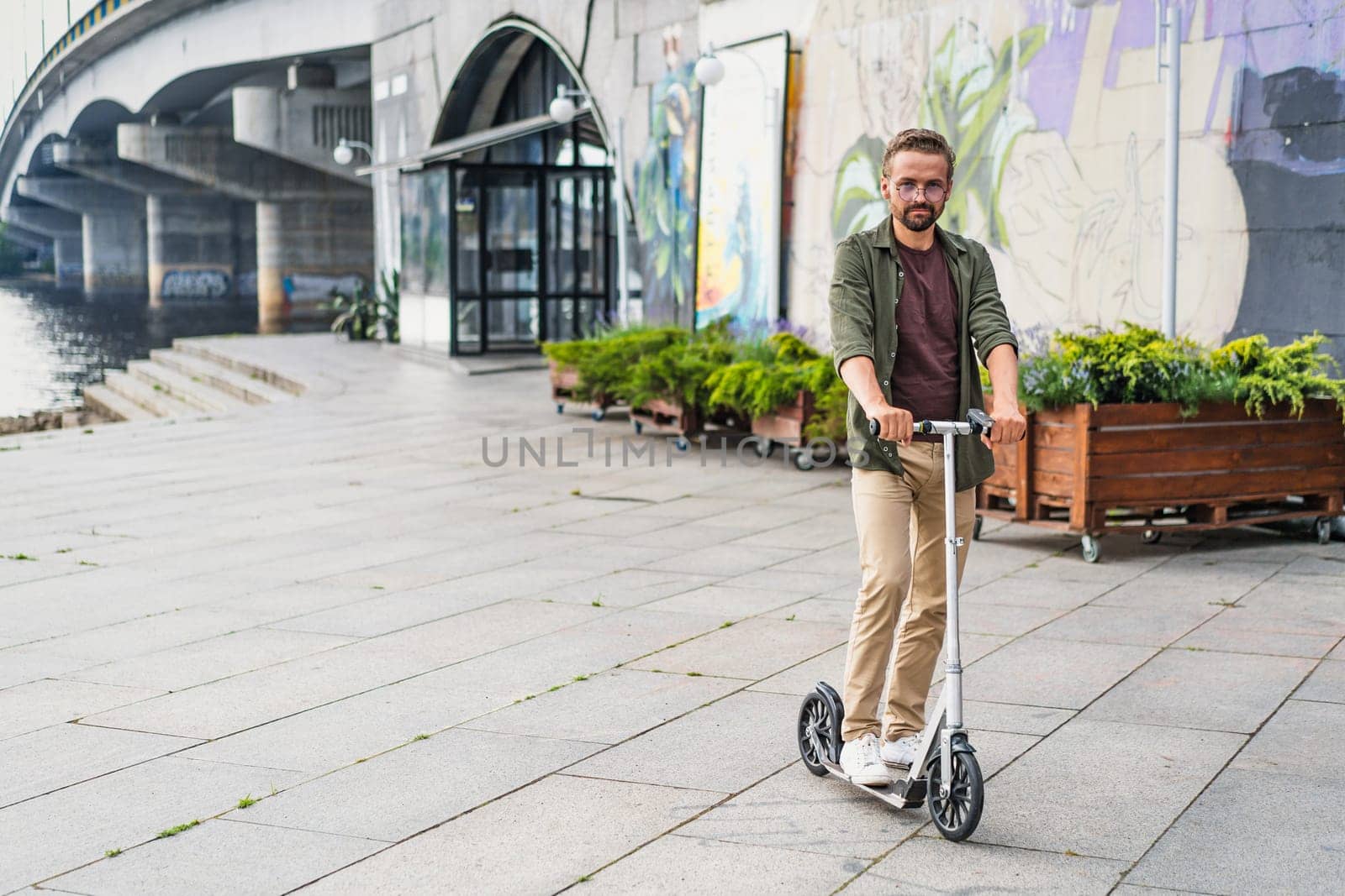 Man riding a scooter as he commutes home from work. The concept of a new city vehicle is embodied by this eco-friendly and efficient mode of transportation. With the urban cityscape in the background. by LipikStockMedia