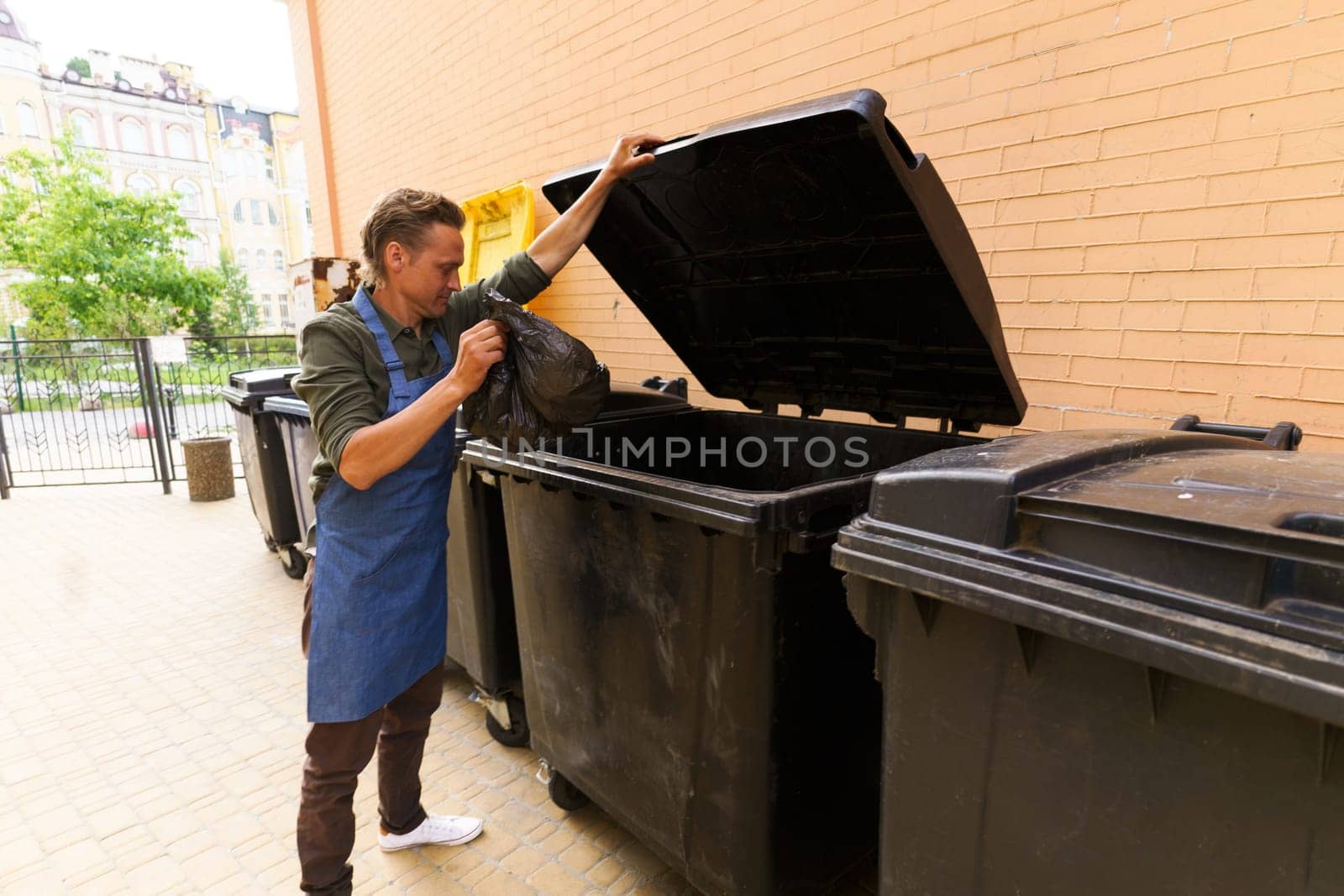 Waiter or kitchen worker responsibly disposing of garbage in pocket and placing it in city trash bin located in back yard. Focus on maintaining cleanliness and proper waste management in urban environment. . High quality photo