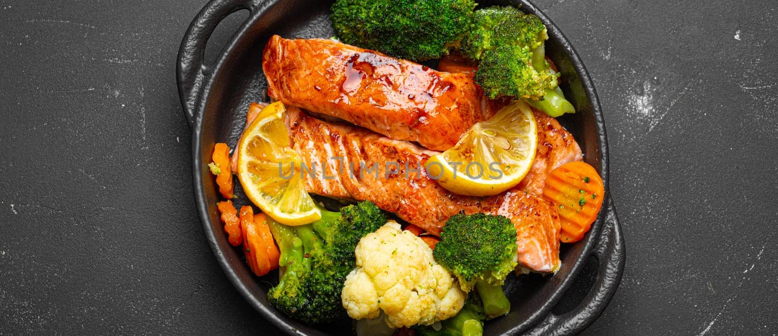Healthy baked fish salmon steaks, broccoli, cauliflower, carrot in cast iron casserole bowl on black dark stone background. Cooking a delicious low carb dinner, healthy nutrition by its_al_dente