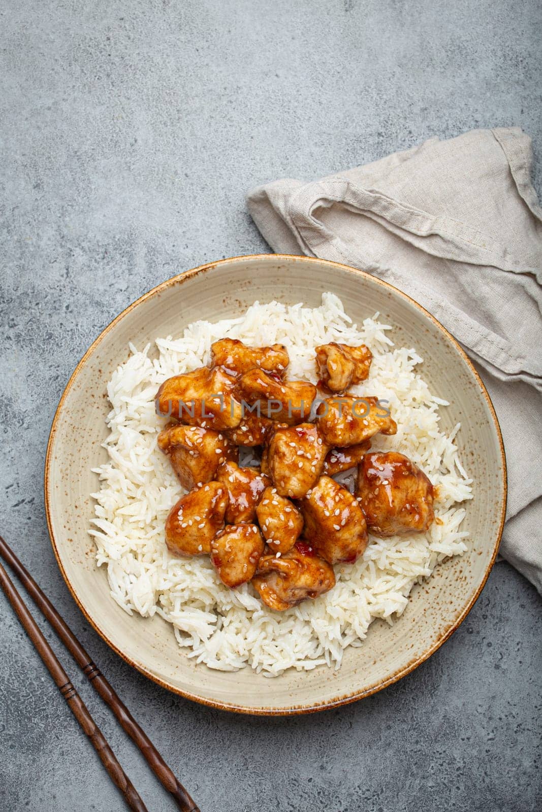 Chinese sweet and sour sticky chicken with sesame seeds and rice on ceramic plate with chopsticks top view on gray rustic stone background, traditional dish of China