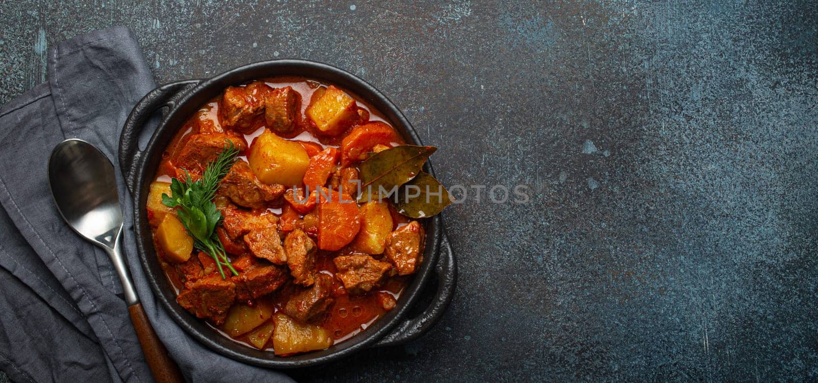 Beef meat stew with potatoes, carrot and delicious gravy in black casserole pot with bay leaves and fresh green herbs with spoon on dark rustic concrete background from above, space for text. by its_al_dente
