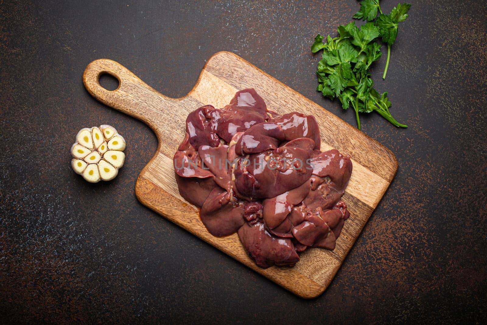 Raw chicken liver on wooden cutting board top view on dark rustic concrete background kitchen table with parsley and garlic. Healthy food ingredient, source of iron, folate, vitamins and minerals