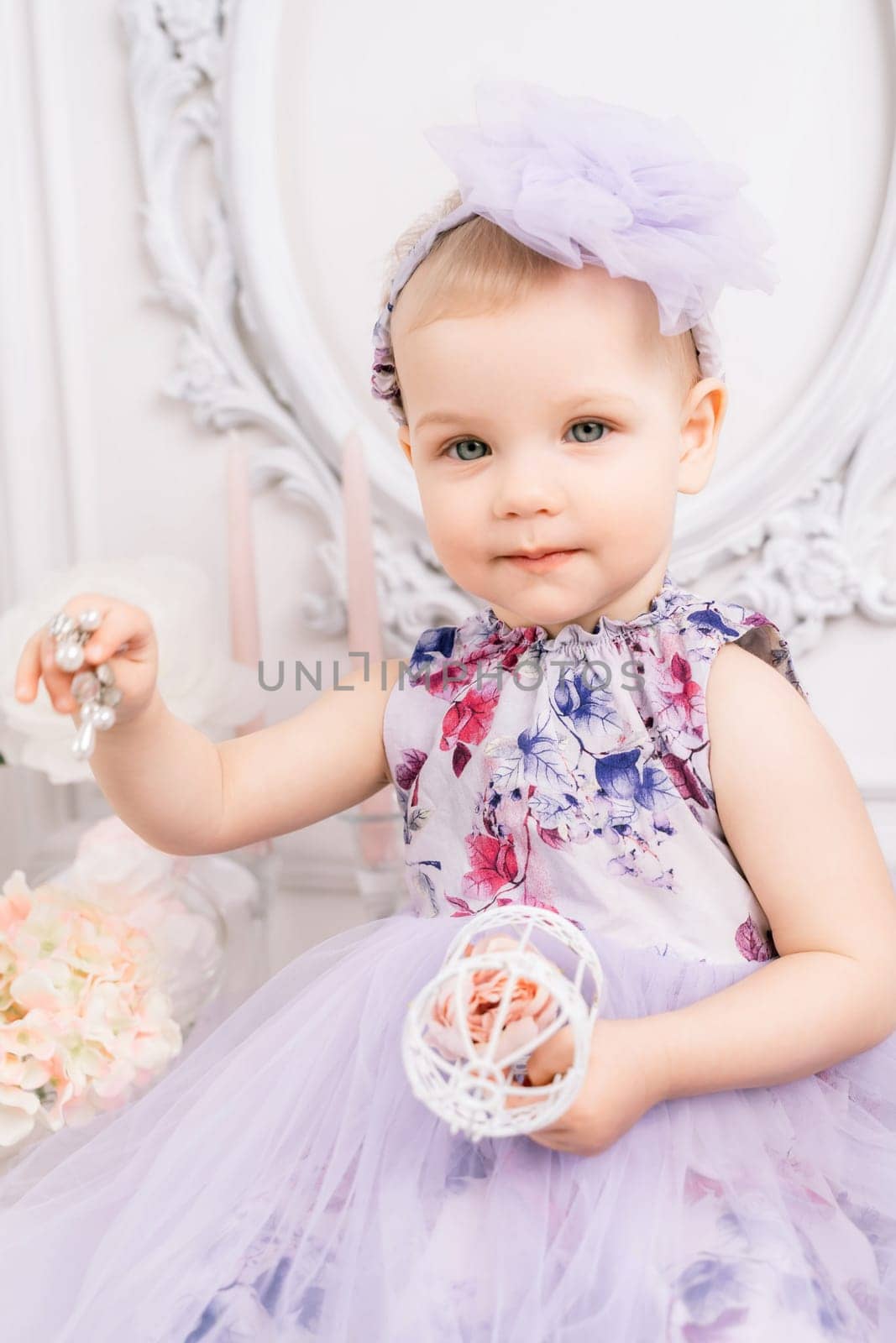 Baby girl elegant dress. A one-year-old girl in a puffy dress and a cute bow poses against the backdrop of a bright room with a dressing table and flowers