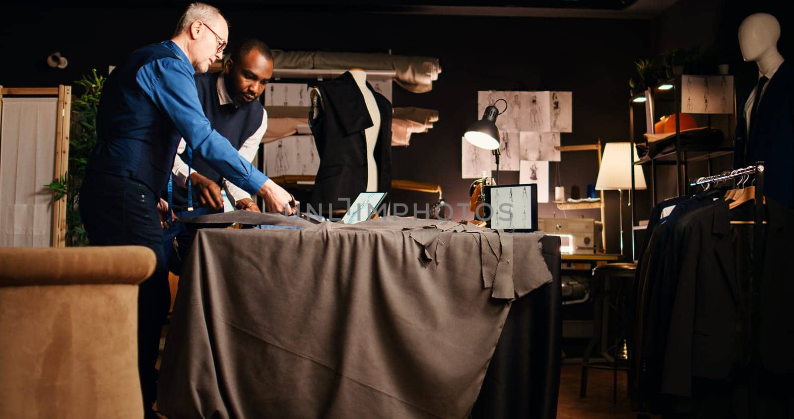 Craftsman and student work on clothes, cutting material for tailoring process. Senior tailor teaching apprentice sewing and handcrafting skills, manufacture fashion items. Handheld shot.