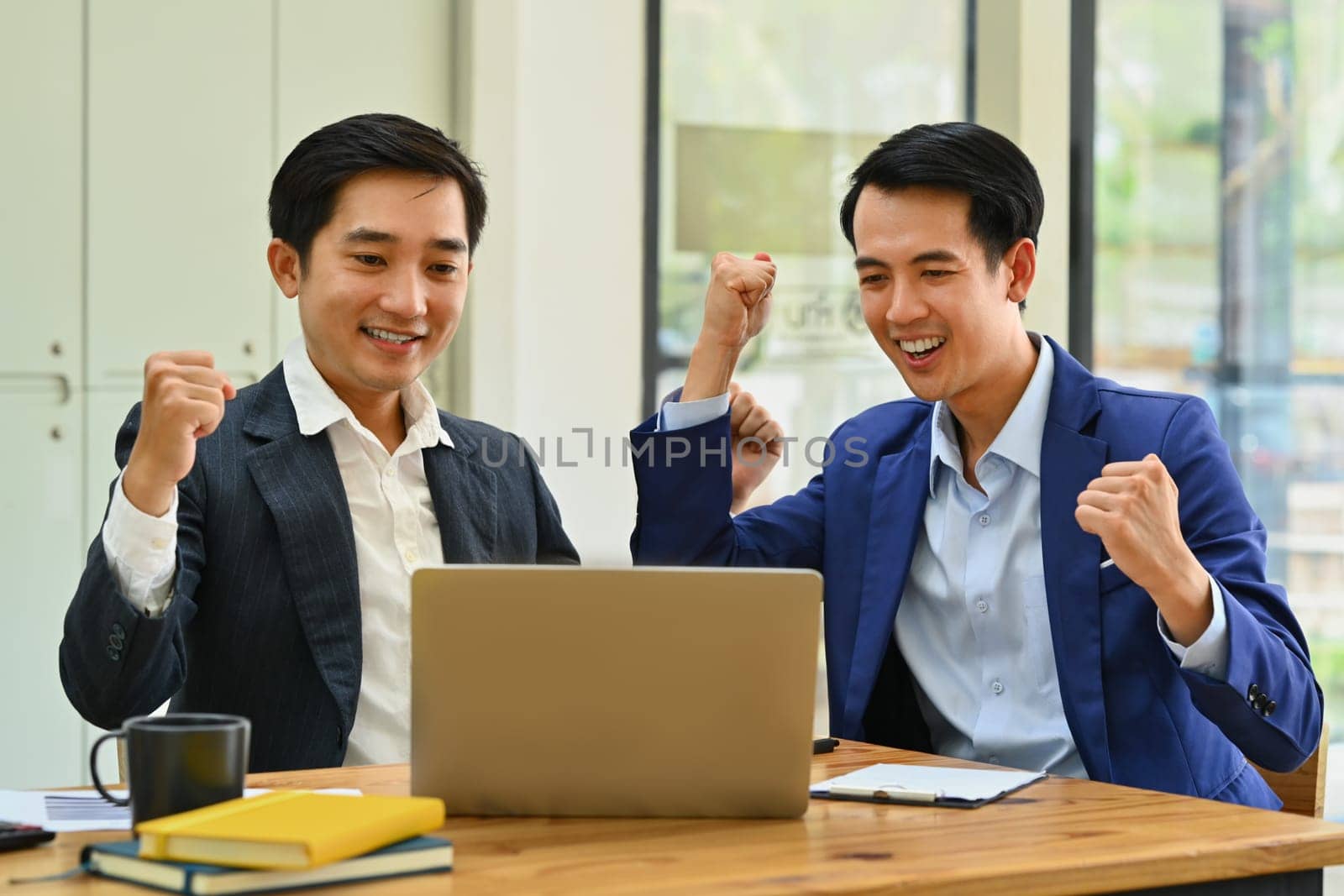 Happy business people looking at laptop screen excited by good news online, celebrating success raising hands up.