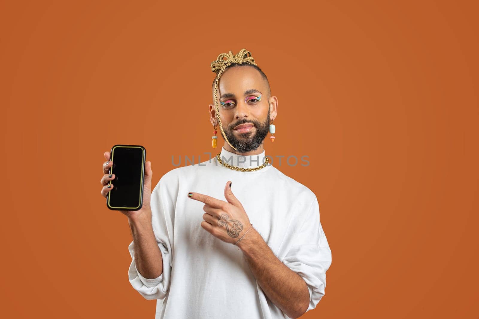 Young smiling african-american gay man wear sweatshirt hold in one hand mobile phone pointing on smartphone screen by finger isolated on orange background studio portrait. Lgbtq pride concept