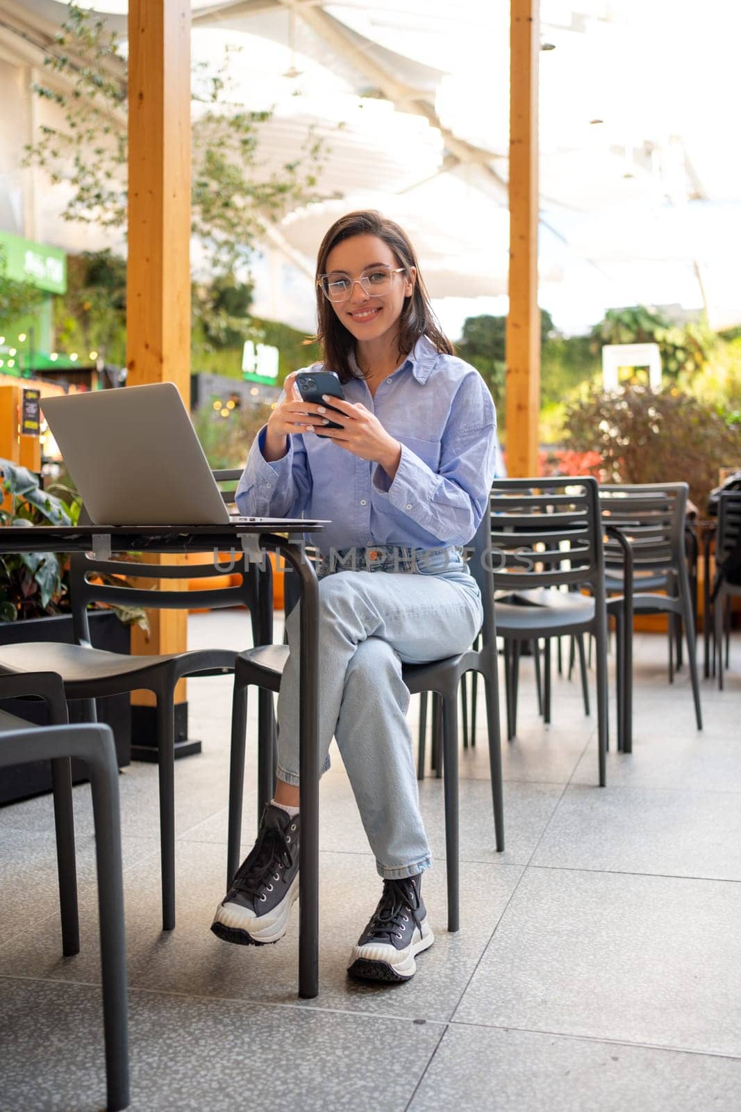 Freelance worker doing remote work using smartphone and laptop, looking at camera and smile. Female freelancer with mobile phone in outdoor cafe. Full length vertical shoot
