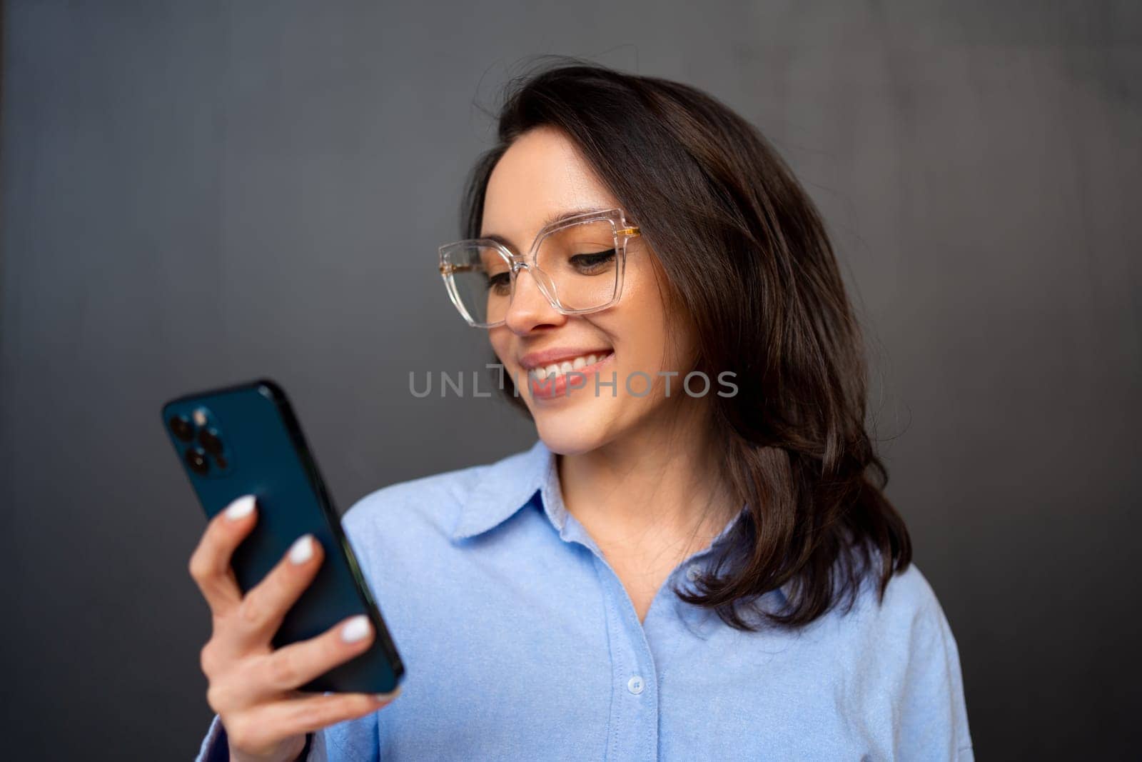Portrait smiling woman holding smartphone in hand looking on mobile phone screen, wear glasses, types text message on mobile phone, enjoys online communication, on black studio background.