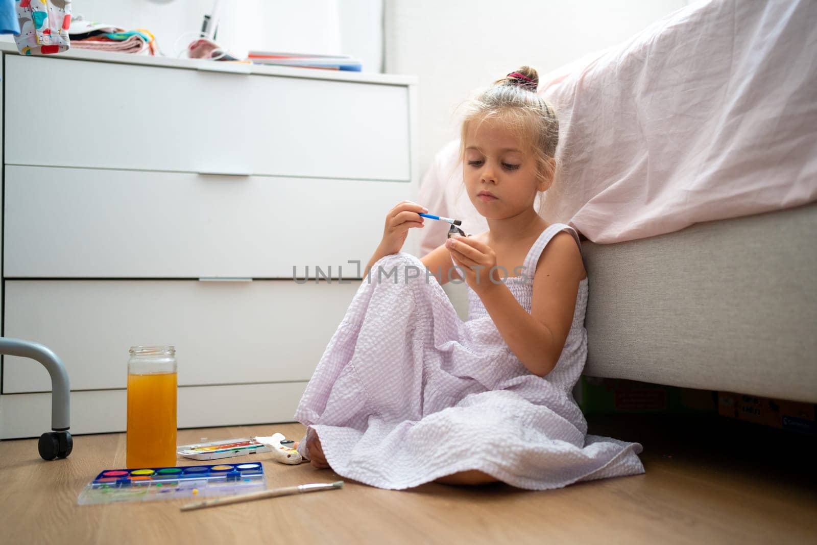 Caucasian child girl is painting watercolor paint on small ceramic figure with fun. Kids arts and crafts creative activity class at home. Little girl make ceramic decor sitting floor