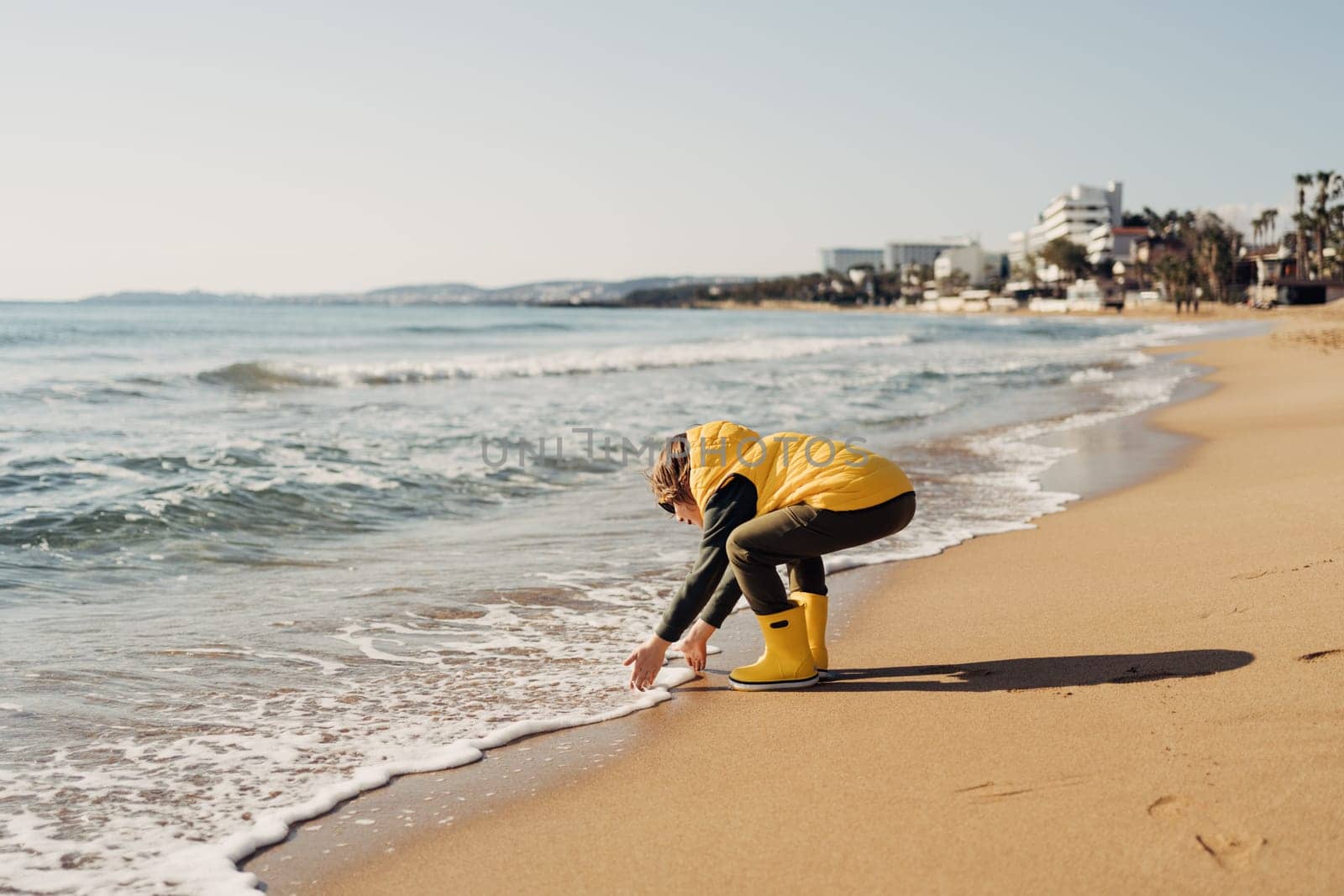 Boy in yellow rubber boots playing with water and sand at the beach. School kid touching water at autumn winter sea. Child having fun with waves at the shore. Spring Holiday vacation concept by Ostanina