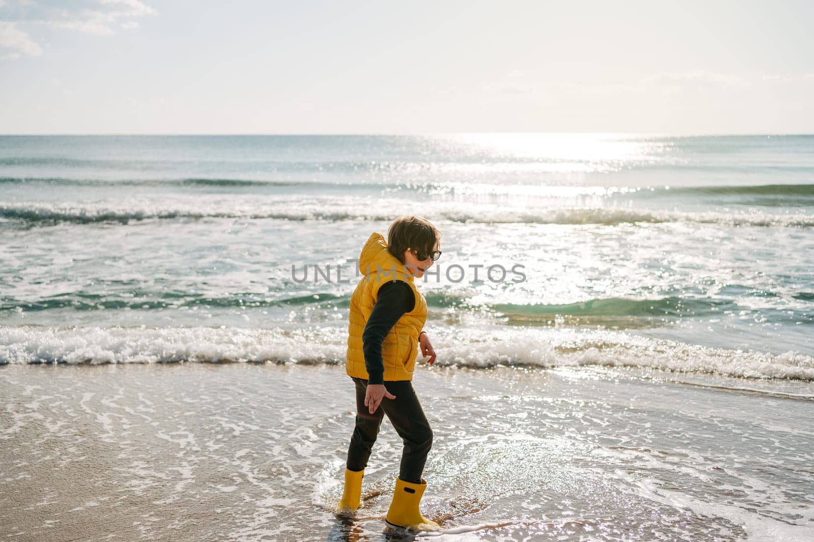 Boy in yellow rubber boots playing with sand at the beach. School kid touching water at autumn winter sea. Child having fun and jumping with waves at the shore. Spring Holiday vacation concept by Ostanina