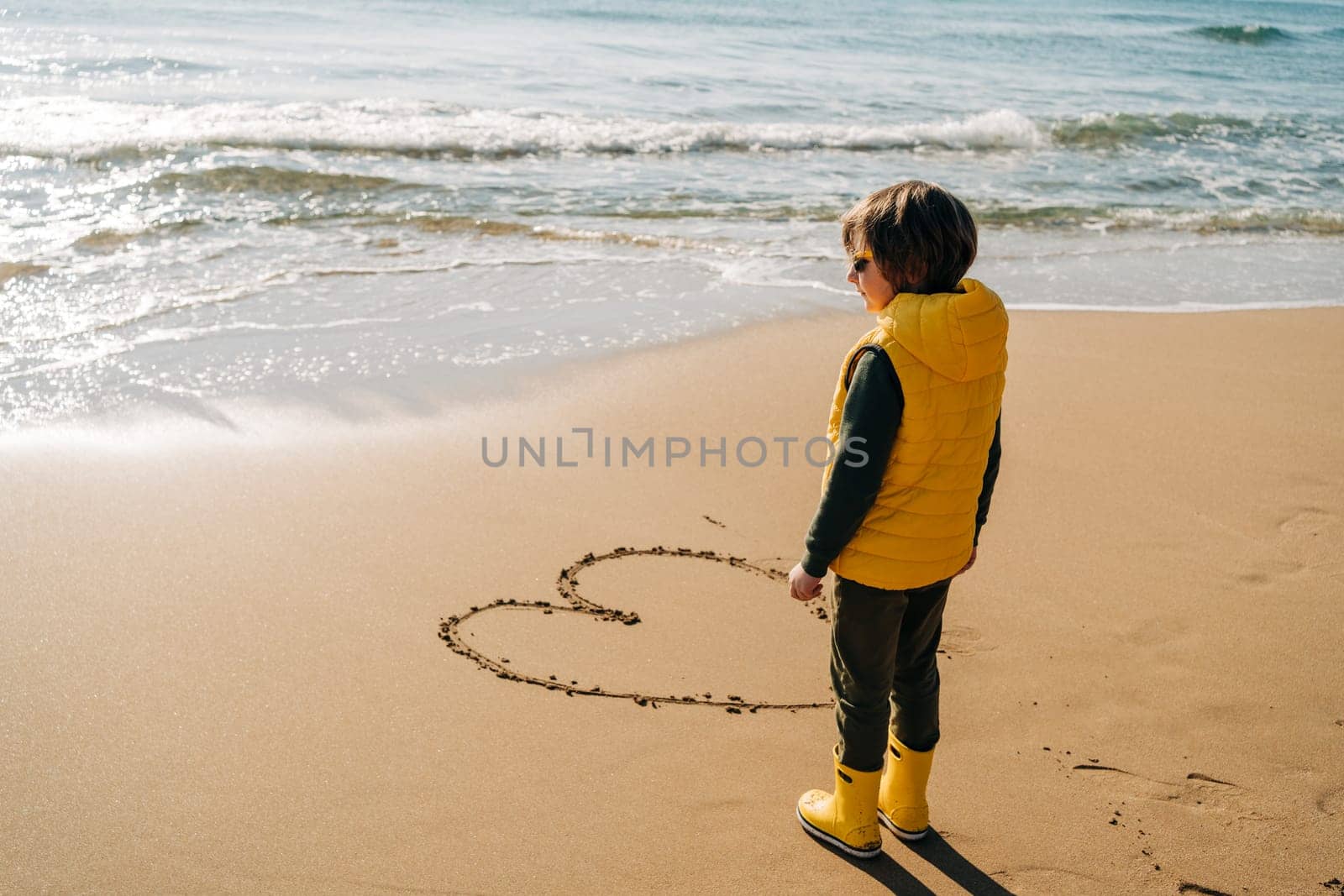 Boy in yellow rubber boots drawing heart shape on sand at the beach. School kid touching water at autumn winter sea. Child having fun with waves at the shore. Spring Holiday vacation concept.