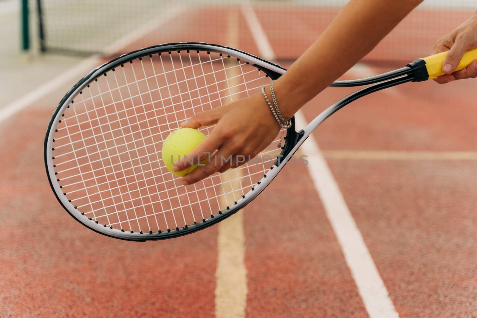 Closeup picture of attractive sporty woman hands holding yellow tennis ball and racket with tennis net and synthetic hard tennis court on the background.