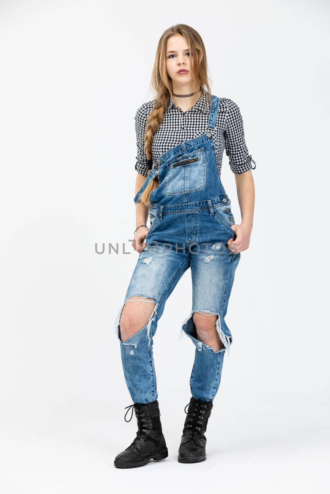 A young girl poses for a photo in a standing position., The girl is wearing a checked shirt and rubbed jeans. The girl is wearing black high top shoes. She is styled in a braid and has disheveled hair.
