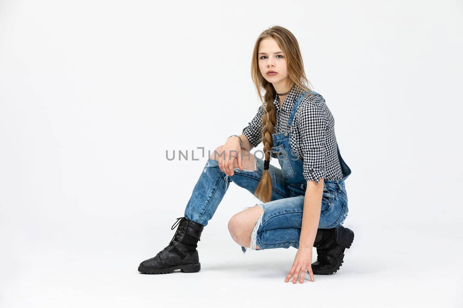 The girl poses against a white background. The woman stays in a crouching position. She is leaning against the ground with one hand. The girl is combed in a thick braid and her hair is in a messy style. She is wearing a checked shirt.