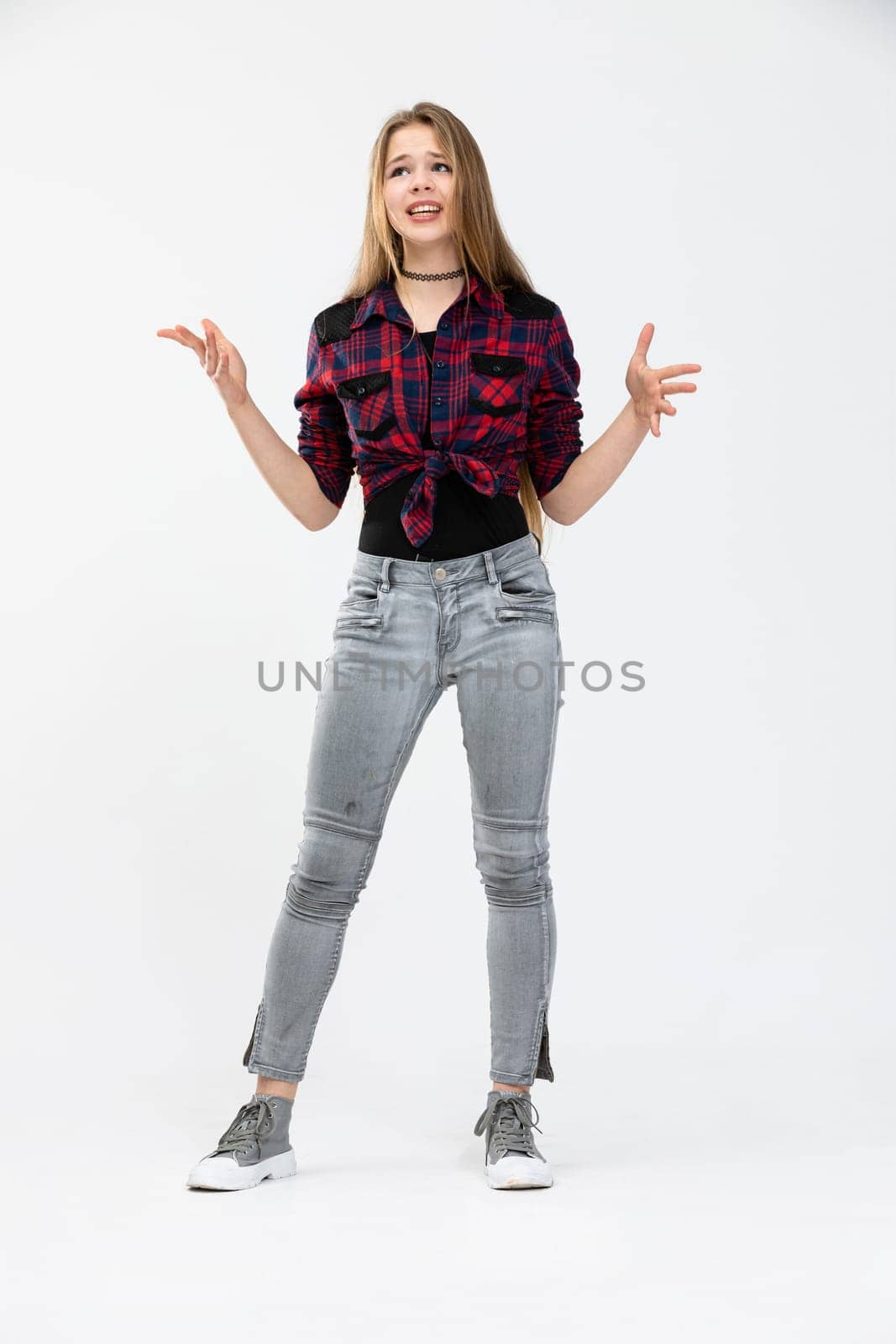 The woman is standing and posing against a white background. She is wearing light tight jeans and a krfata shirt tied in a knot. The girl has her arms spread at her sides and poses with a disgruntled expression. Long-haired girl with loose hair.