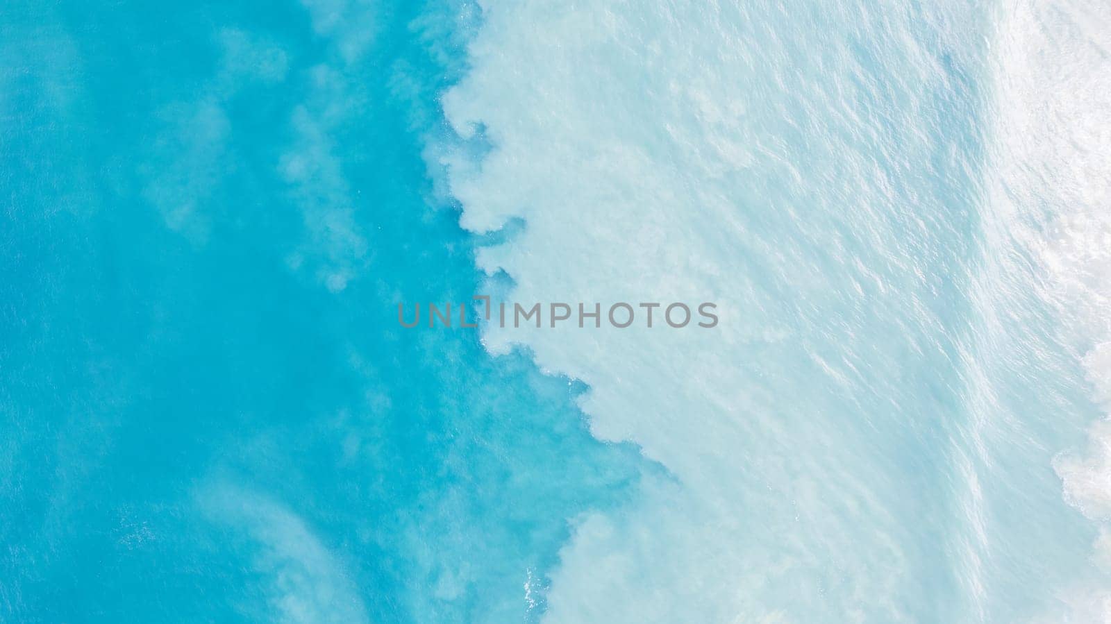 Top view of light blue water with white sand. Waves raise sand and a mix with turquoise water is obtained. Shadows from clouds on a clear ocean. Clear sea. Gradient of blue. The background photo