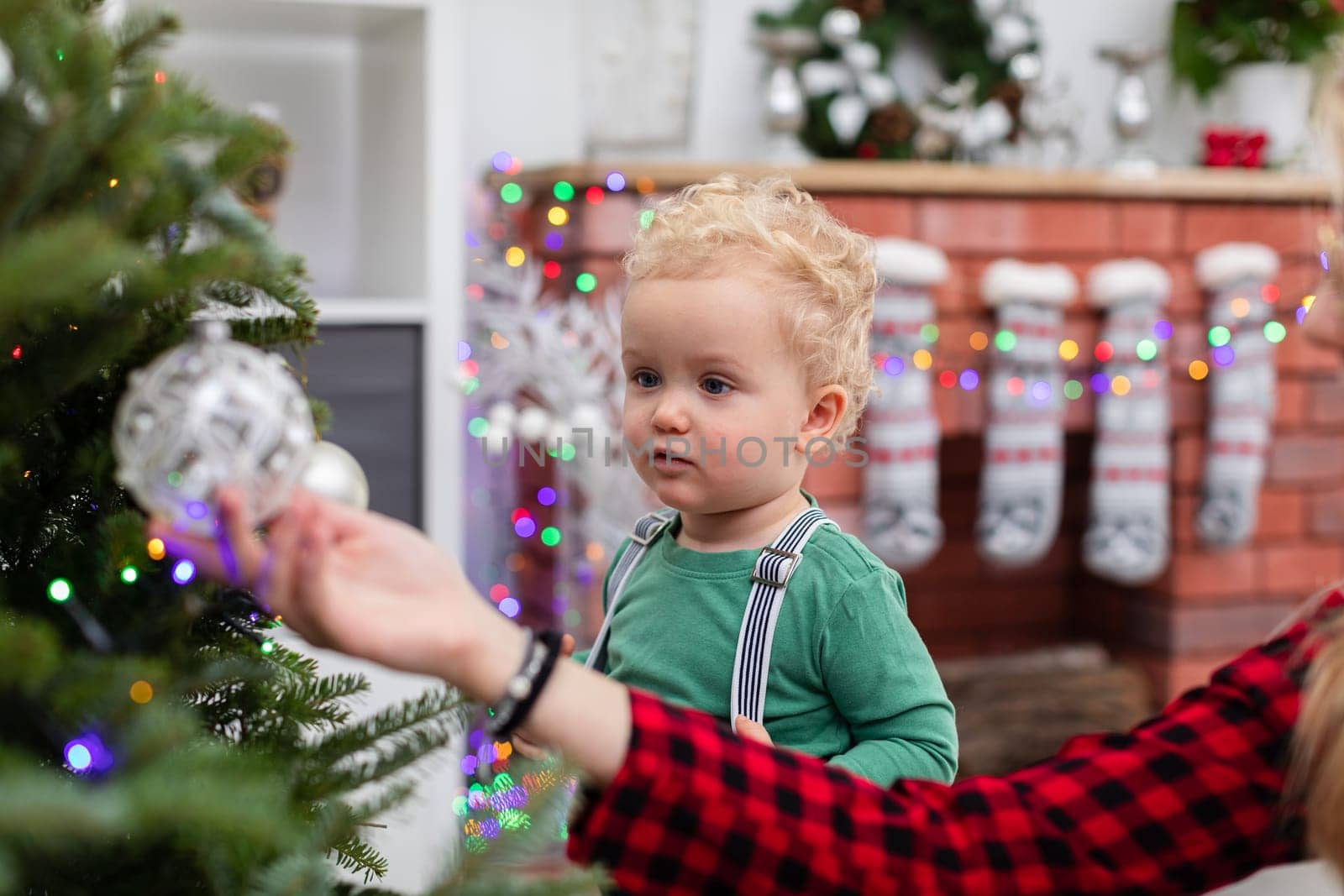 Blurred foreground. In the foreground a woman's hand showing a bauble hanging on a Christmas tree. A little boy stands next to her and looks at the bauble. In the background a fuzzy brick fireplace decorated with colorful lights.