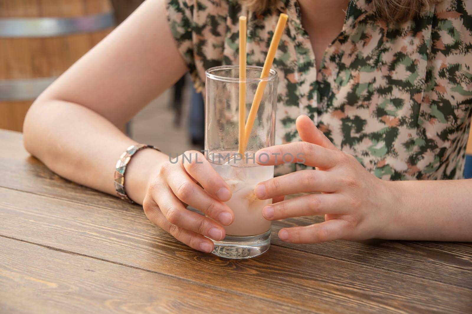 Cropped close-up of unrecognizable young woman holding glass with cold exotic cocktail, sitting at table in street cafe on sunny gold time, woman drinking cool lemonade. High quality photo