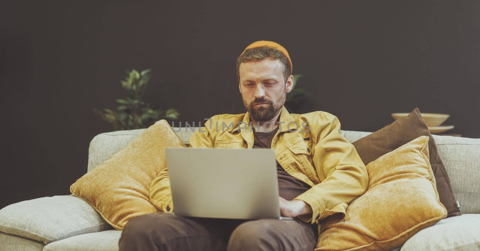 Man sitting on couch with a laptop, engaged in work or other digital activities. Relaxed and comfortable setting of couch suggests that man is in home office or cozy indoor environment. by LipikStockMedia