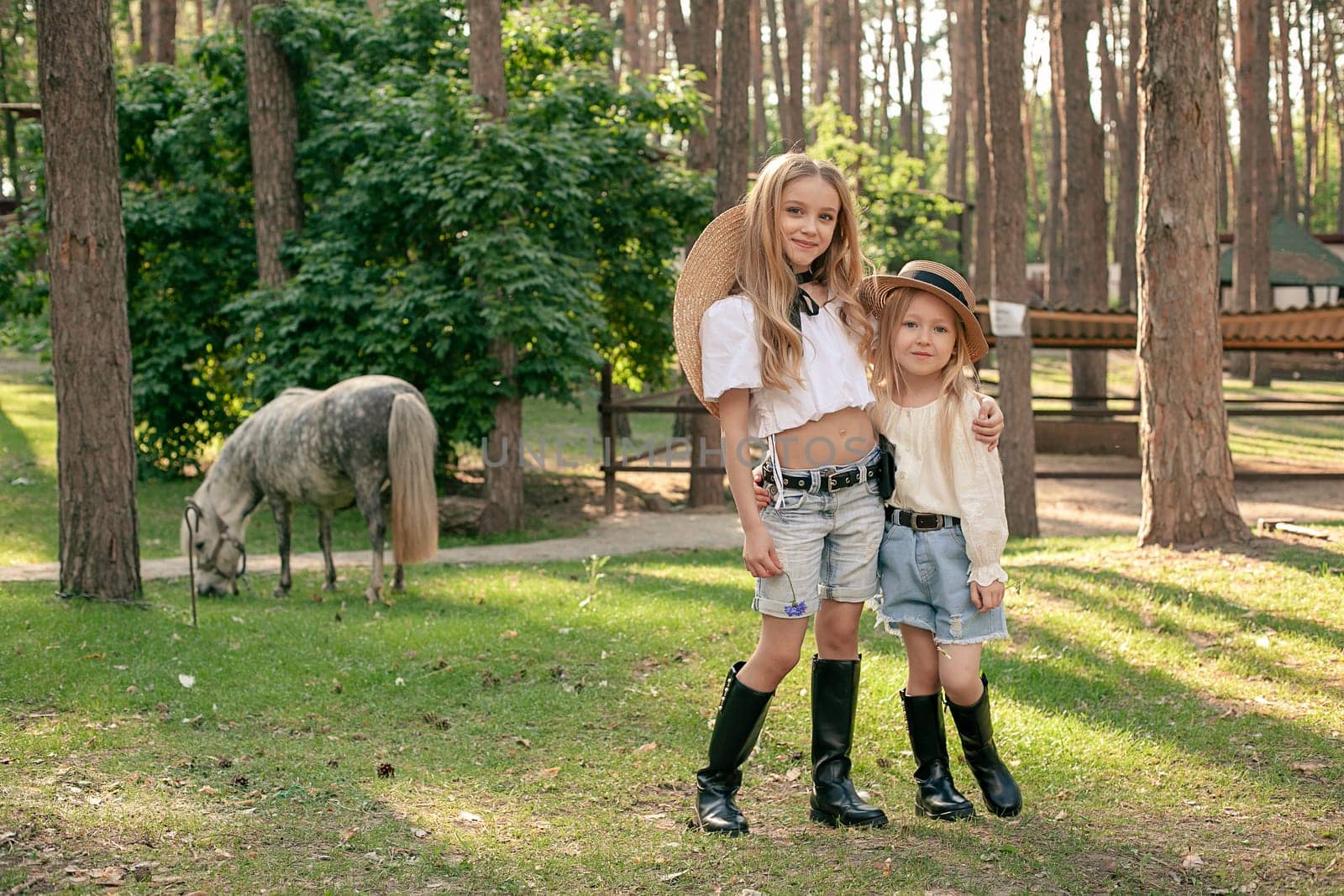 Two happy tween girls standing on green lawn on pine park on background of grazing dapple gray pony. Portrait of two smiling sisters hugging while walking. Sisterly affection concept