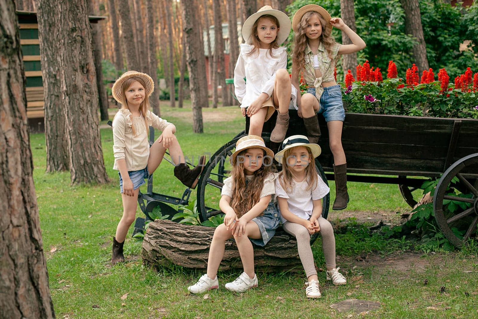 Happy carefree tween girls sitting on old wooden cart decorated as flower bed with blooming red salvia in courtyard of country estate located in pine forest on sunny day