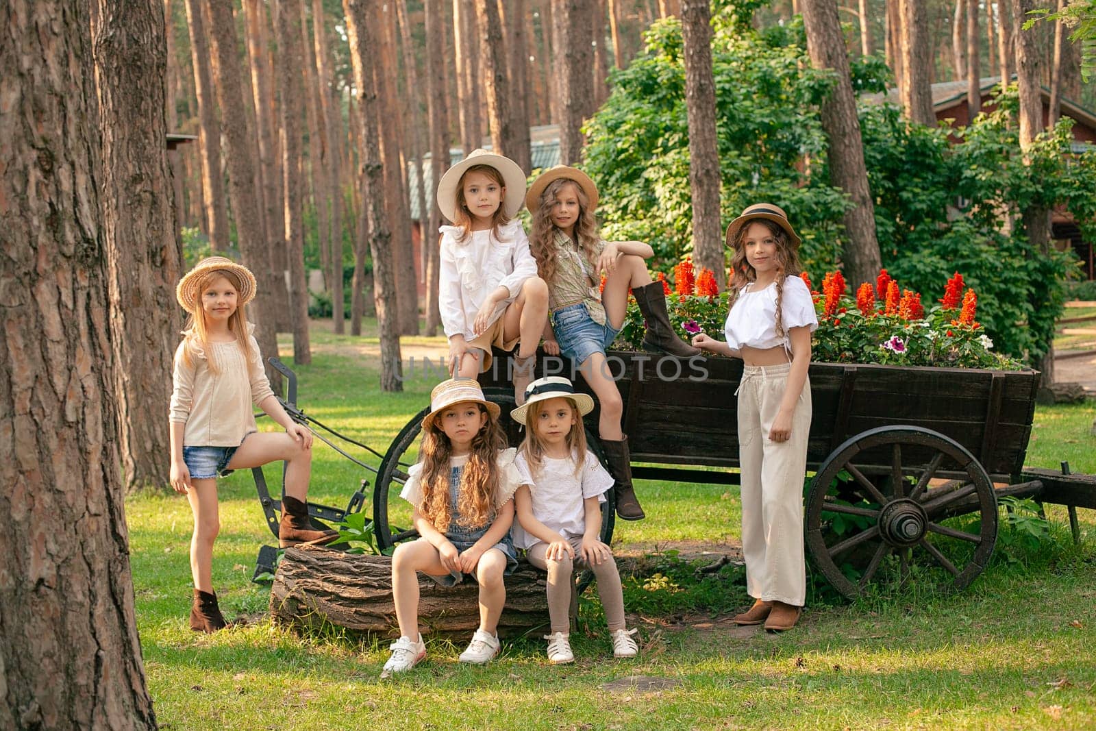 Cheerful preteen girls enjoying summer vacation in country estate, posing together outdoors near old rustic wooden cart used as designer flower bed with blooming red salvia