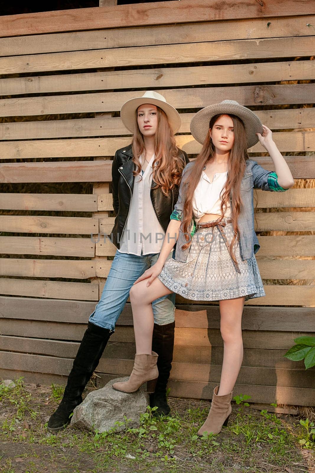 Two attractive long-haired teen girls in rustic style clothes and wide-brimmed hats posing outdoors against wooden boards background, looking confidently at camera