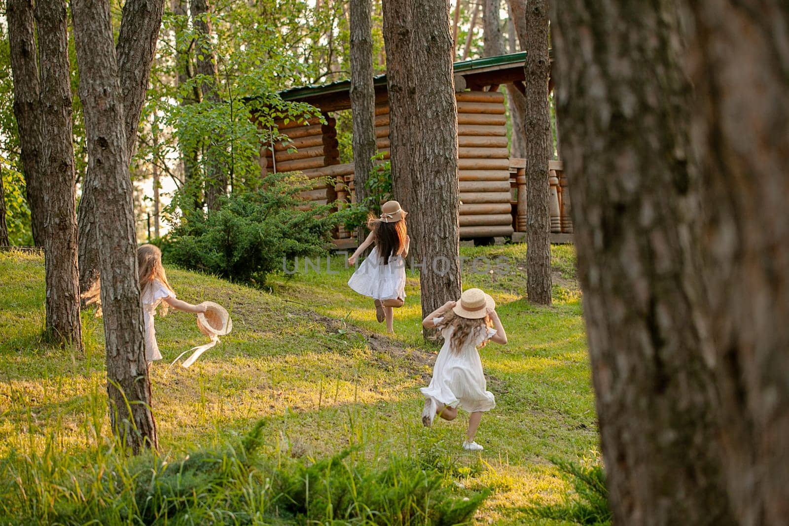 Rear view of preteen girls in light dresses and wicker hats having fun in country estate on sunny summer day, playing tag running among tall trees on green lawn