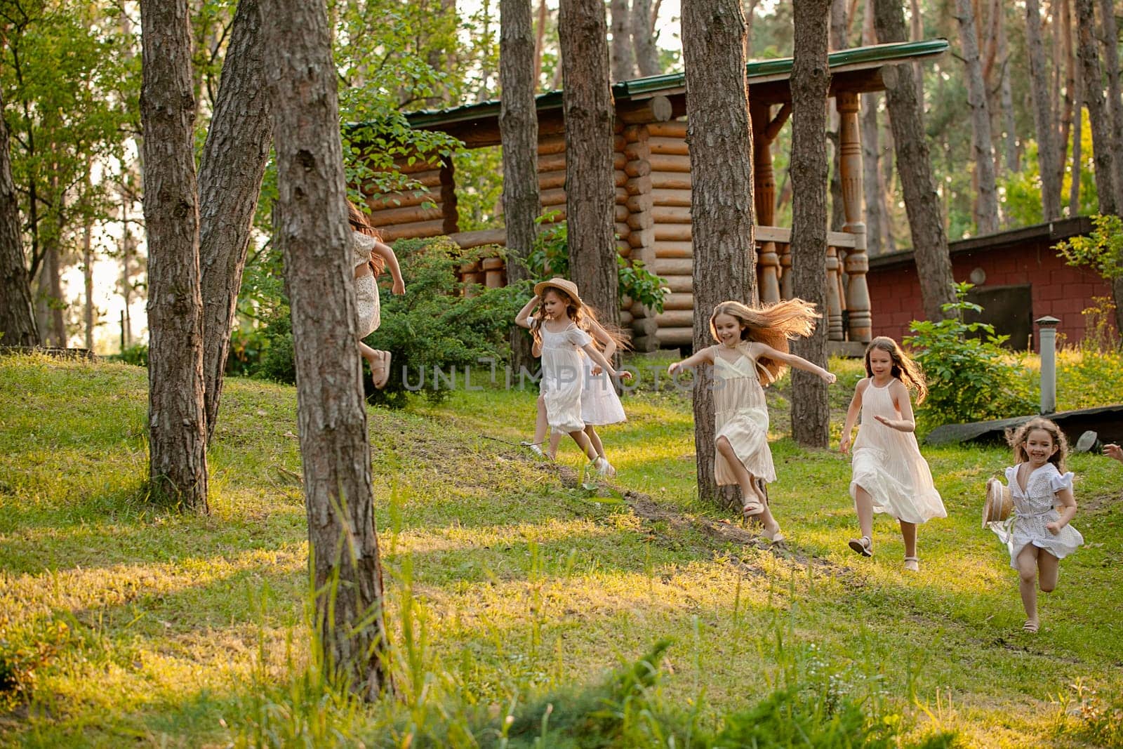 Group of happy preteen girls in light dresses having fun in country estate on sunny summer day, playing tag running among tall trees on green lawn