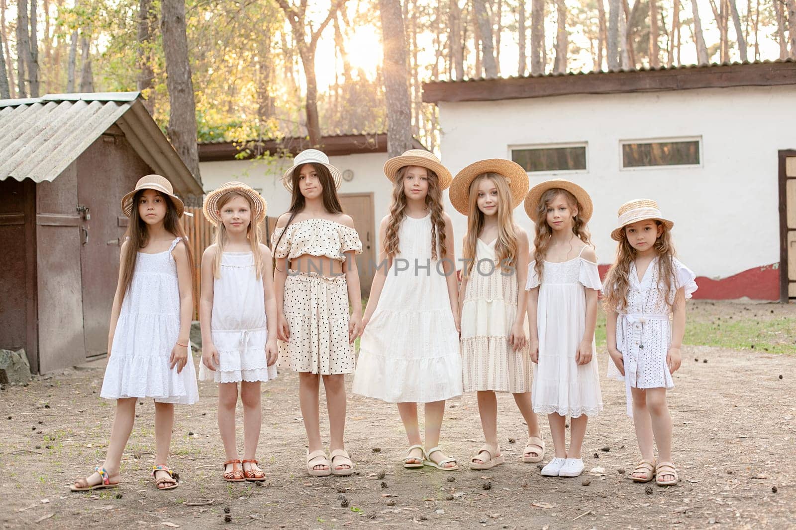 Trendy stylish preteen girls in similar light dresses and wicker hats with brim standing in backyard of country house located in forest against backdrop of outbuildings and tall trees in summer