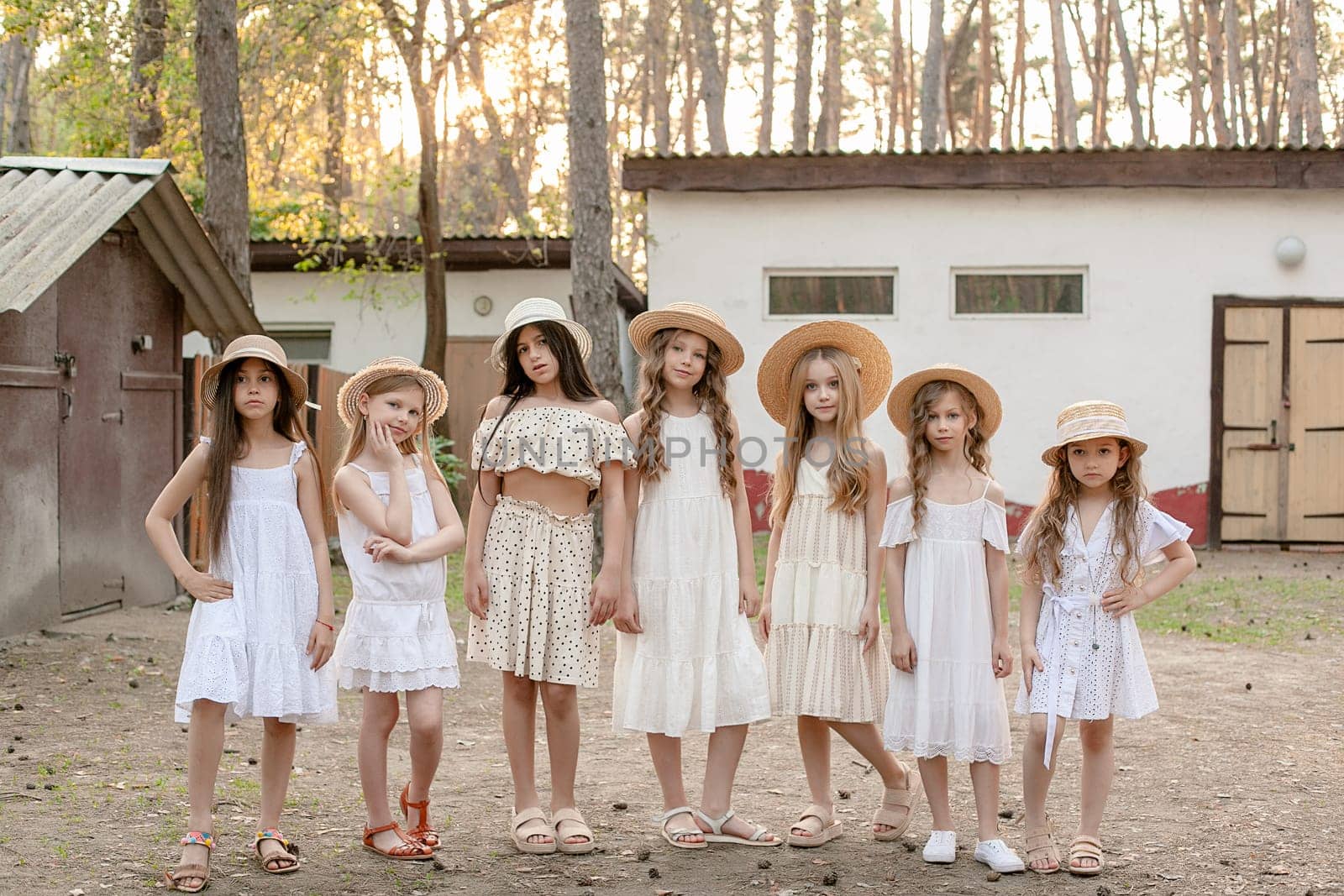 Smiling friendly tween girls in light dresses and straw sun hats standing on courtyard of country house located in pine forest on sunny summer day, enjoying summer vacation